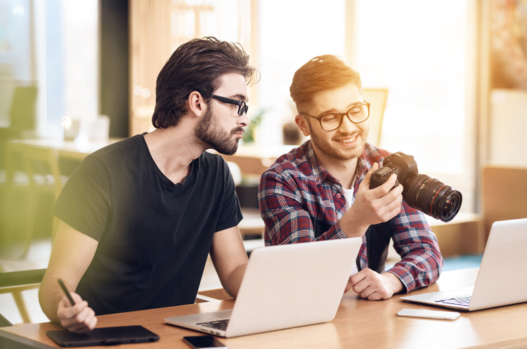 Two men working at laptops while one holds a camera